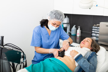 Qualified woman cosmetologist performing ultrasound cavitation body procedure for elderly female client at aesthetic cosmetology clinic.