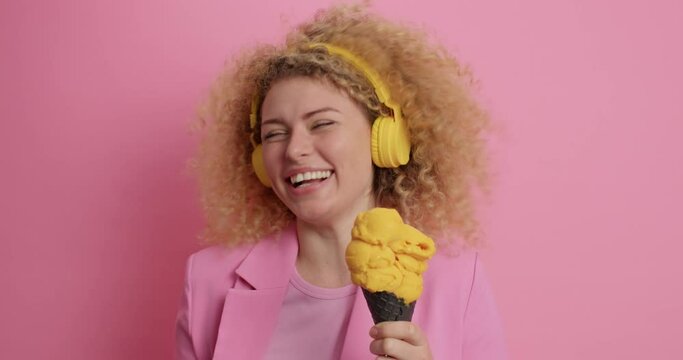 Summer food concept. Smiling pleasant looking curly woman has fun enjoys eating delicious yellow ice cream listens favorite music via headphones grins at camera poses against pink background