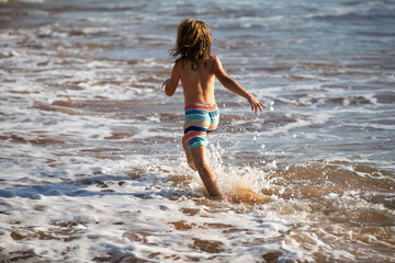 Fototapeta premium Back view of kid boy have fun on tropical sea beach. Funny child run with splashes by water pool along surf edge. Kids activity on summer holiday.