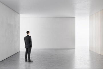 Businessman in bright concrete interior with empty wall looking at mockup place for your...
