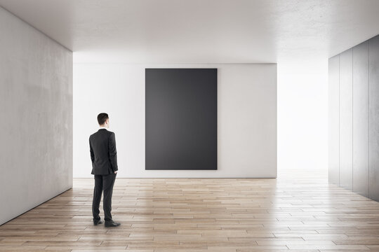 Businessman in modern concrete interior with empty poster looking at mockup place for your advertisement.