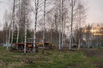 Old red and yellow tractors stand among the trees in the open air on a driverless farm. Agricultural machinery