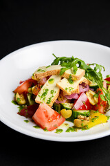 Vegetable salad with cheese, fresh romatoes and cucumbers, green olives served in a white plate over black background.