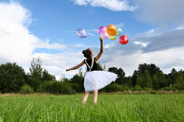 happy girl in beautiful dress holds colorful balloons in summer meadow against  background of blue sky and clouds, has fun and throws them up. The concept of  festive mood, birthday