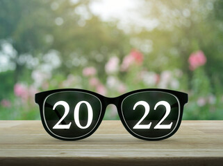 2022 white text with black eye glasses on wooden table over blur pink flower and tree in garden, Business vision happy new year 2022 cover concept