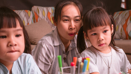 Obraz premium Happy mother and daughters learned to draw through the laptop computer in the living room. Mother helping the girls to draw a picture on paper. Home school concept. Kindergarten online classes.