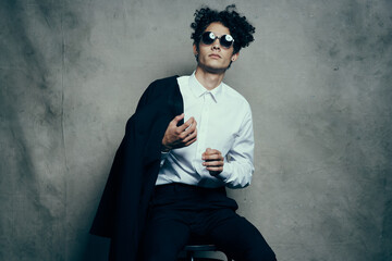  guy in a suit sits on a chair indoors and glasses on his face, a jacket in his hand