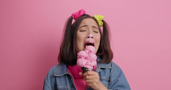 Dejected Asian woman sobs and cries has troubles in life licks ice cream tries to sooth herself smeared with sweet dessert wears denim jacket isolated over pink background. Upset teenager with gelato
