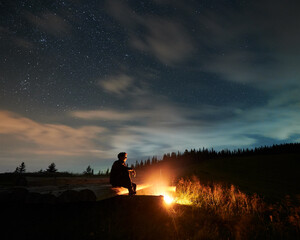 Fototapeta na wymiar Tourist man sitting near cozy bonfire and relaxing on fresh air. Concept of good weather and process enjoying incredible starry sky at night.