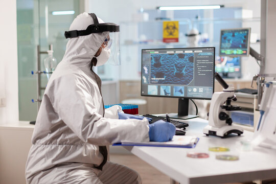 Scientist in ppe suit taking notes on clipboard working on virus treatment. Examining vaccine evolution using high tech technology and chemistry tools for scientific research virus development.