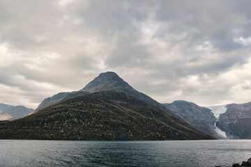 Huge mountain in a Norwegian fjord near a glacier. Dramatic view of a volcano-shaped hill under a cloudy sky.