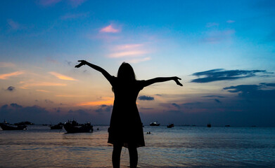 Fototapeta premium Back view silhouette young girl asian with happy and peacful pose on the beach at sunset. Beautiful blonde woman with long hair relaxing at the ocean. Concept of happy, peaceful, relax