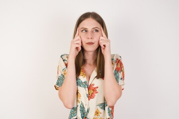 Dreamy Caucasian female with thoughtful expression, looks away, keeps hand near face, thinks about something pleasant, poses against white wall.