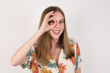 Beautiful young blonde woman with happy face smiling doing ok sign with hand on eye looking through finger. Wearing casual clothes and standing.