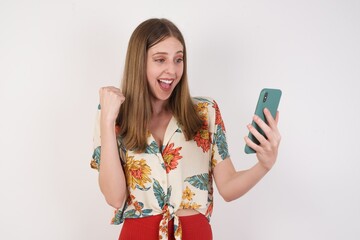 Portrait of a happy cheerful woman celebrating success while standing and looking at mobile phone isolated over white bright background.