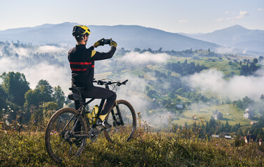 Back view of young man in cycling suit sitting on bicycle and talking mountain photo with modern...
