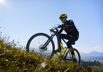 Smiling man in safety helmet and glasses cycling uphill on sunny day with blue sky on background. Male bicyclist in cycling suit climbing uphill on mountain bike. Concept of sport and active leisure.