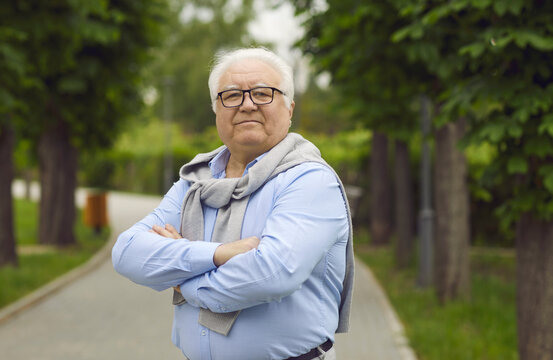 Portrait of confident senior citizen. Elderly man in glasses and shirt standing on park pathway and looking at camera. White-haired old grandfather spending time in green park in spring or summer