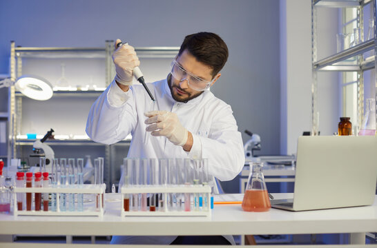 Serious concentrated young male scientist, pharma chemist, biotech company employee or student in coat and lab glasses working alone in science laboratory transferring liquid in sterile glass tube