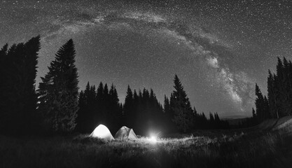 Panoramic view of night camping in valley with large pine trees. Burning campfire and illuminated tourist tents under bright starry sky with Milky Way. Black and white image.