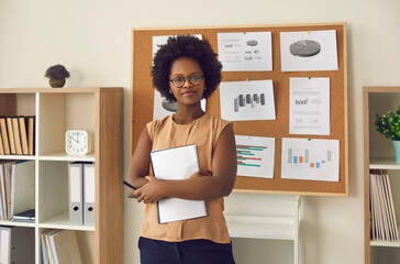 Portrait of smart confident smiling young african american woman executive manager speech presenter with arm folded standing holding notebook near blackboard with project result report in office