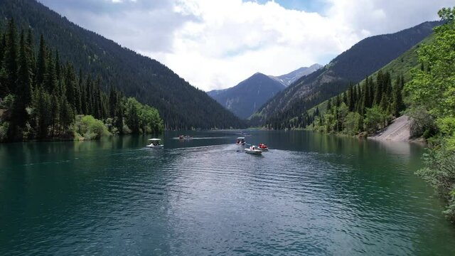 Kolsai mountain lake and green forest. Top view. People swim in boats and catamarans. They rest on the green meadows. The sun's rays shine on water. Clear water and tall coniferous trees. Kazakhstan