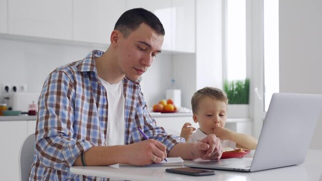 Young dad sitting at home in kitchen with kid of 3-4 years old, working learning on laptop watching online classes making notes. Parent distance work at home office and child care concept. - Powered by Adobe