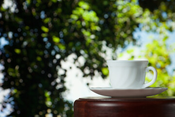 White cup of coffee or tea on a wooden table over blurred tree with sun lighting
