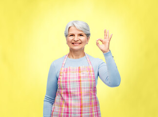 cooking, culinary and old people concept - portrait of smiling senior woman in kitchen apron showing ok hand sign over illuminating yellow background