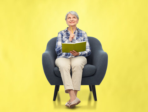 inspiration, planning and old people concept - smiling senior woman sitting in modern armchair and writing to diary over illuminating yellow background