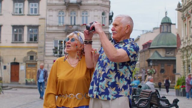 Senior old stylish tourists man woman walking, taking photos on old camera at summer city center Lviv, Ukraine. Elderly grandmother grandfather enjoying conversation, traveling together. Mature family