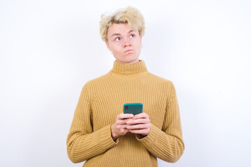 Portrait of Young handsome Caucasian blond man standing against white background with dreamy look, thinking while holding smartphone. Tries to write up a message.