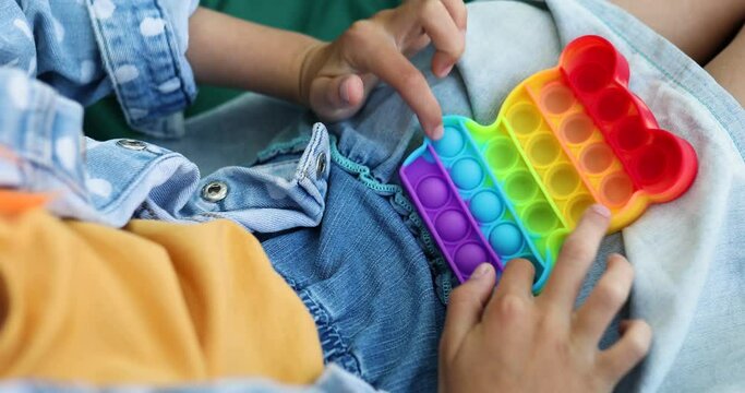 A little girl the on hanging chair outdoors play pop it, kid hands playing bubbles of a rainbow destressing toy, fidget toy in backyard of house on a sunny summer day, summer time vacation.