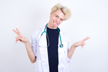 young handsome Caucasian doctor man standing against white background with optimistic smile, showing peace or victory gesture with both hands, looking friendly. V sign.