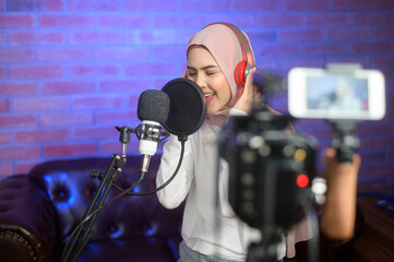 Young smiling muslim female singer wearing headphones with a microphone while recording song in a...
