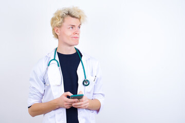 Portrait of young handsome Caucasian doctor man standing against white background with dreamy look, thinking while holding smartphone. Tries to write up a message.