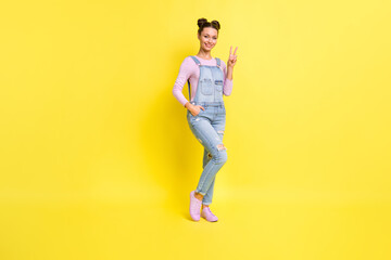 Fototapeta na wymiar Photo of charming young girl posing show v-sign shiny toothy smile wear denim overall shoes isolated yellow background