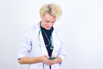 Smiling young handsome Caucasian doctor man standing against white wall using cell phone, messaging, being happy to text with friends, looking at smartphone. Modern technologies and communication.