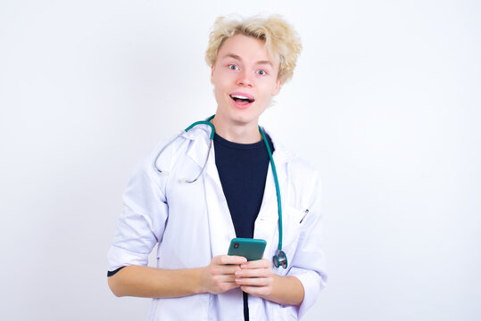 Smiling young handsome Caucasian doctor man standing against white background friendly and happily holding mobile phone taking selfie in mirror.