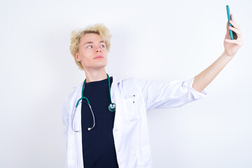 Portrait of a young handsome Caucasian doctor man standing against white background taking a selfie to send it to friends and followers or post it on his social media.