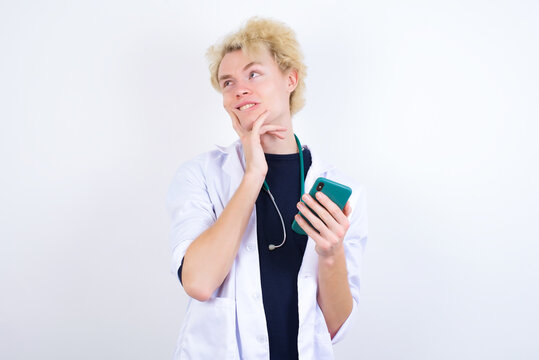 Image of a thinking dreaming young handsome Caucasian doctor man standing against white background using mobile phone and holding hand on face. Taking decisions and social media concept. - Powered by Adobe