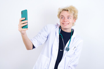 Isolated shot of pleased cheerful young handsome Caucasian doctor man standing against white background, makes selfie with mobile phone. People, technology and leisure concept