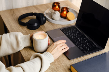 A woman works on a laptop in a cozy environment at home. Freelance girl. Girl in casual clothes. Internet business. Distant work. Girl listens to music on headphones, drinks tea and lights candles