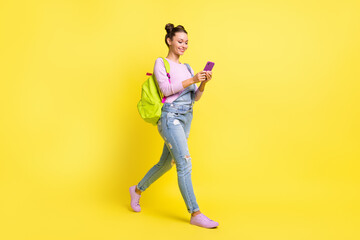 Photo of funny pretty young lady dressed jeans overall backpack typing modern gadget walking isolated yellow color background