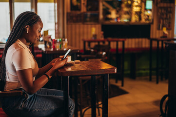 Cheerful african american woman using a smartphone and airpods while sitting in a cafe