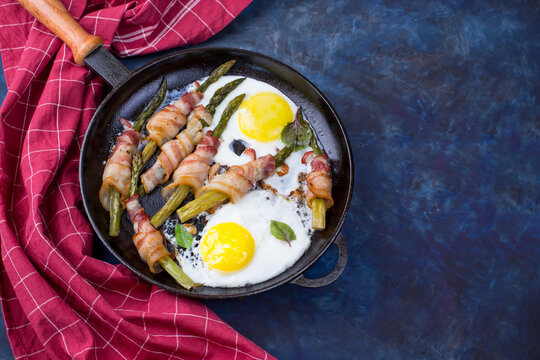 A dish of asparagus, bacon and eggs in a cast-iron skillet on a dark background.