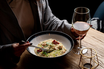 Young attractive woman is eating risotto with a glass of white wine, side view, dark elegant photo with female hands in a frame