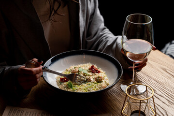 Young attractive woman is eating risotto with a glass of white wine, side view, dark elegant photo with female hands in a frame