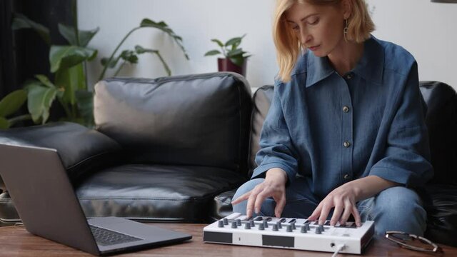 Young woman in casual clothes composing on midi controller in living room, music producer using laptop. Student at online music school, songwriter