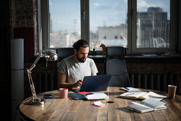 Handsome young brunette male with beard sitting in office at the table working on laptop